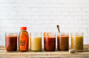 A row of assorted homemade spreads in jars with a bottle of raw honey, against a white brick wall.