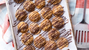 Caramel cookies drizzled with chocolate on a serving tray and a spatula on the side, over a checkered napkin.